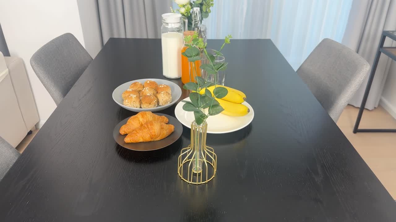 A rotating view of a breakfast table with bananas, croissants, milk, and orange juice in a bright dining room