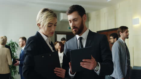 caucasian businesswoman with a folder of documents in hands talking and discussing something with a businessman who holding a tablet during a break in a conference hall