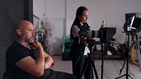 bearded man sits on the chair looking at camera display. long-haired brunette girl taking video on her phone in the studio.