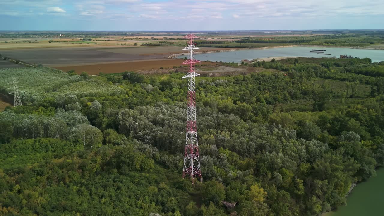 Orbital aerial view as a tall electricity pylon rises above the trees, surrounded by mining lakes and agricultural fields near Szalkszentmárton in Hungary