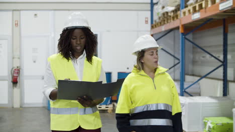 trabajadoras multiétnicas en casco caminando y contando bienes en stock