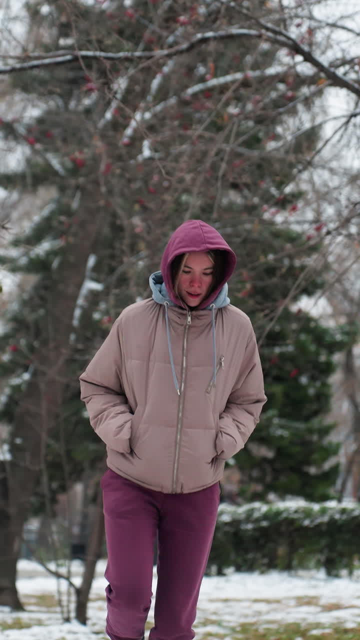 joven salta felizmente en el parque de nieve de invierno, vestida con una sudadera con capucha y abrigo cálido, los árboles cubiertos de nieve crean un fondo escénico, mientras disfruta juguetón del clima frío en un entorno al aire libre sereno