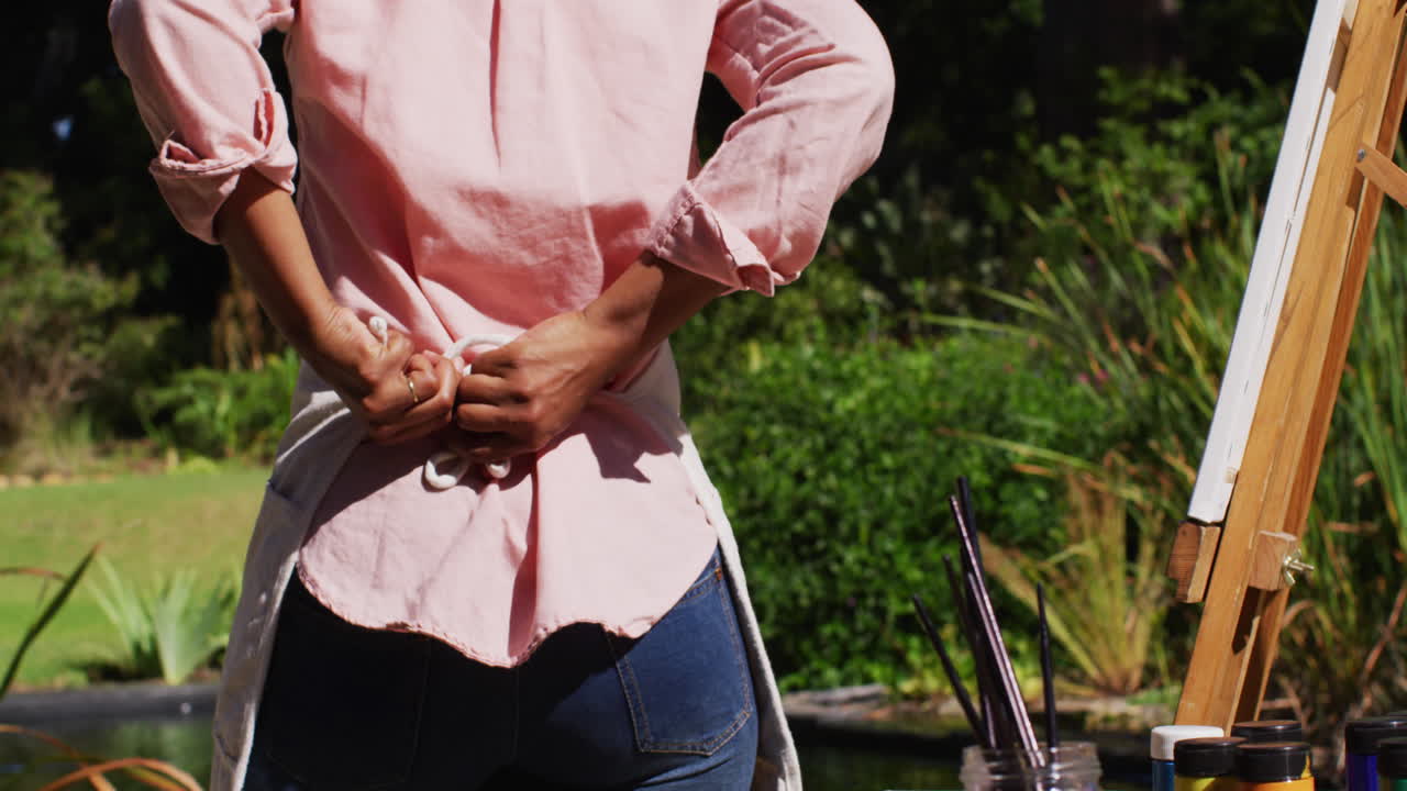 아프리카계 미국인 여성이  ⁇ 은 정원에서 캔버스에 그림을 그리며 애프론을  ⁇ 고 있습니다.