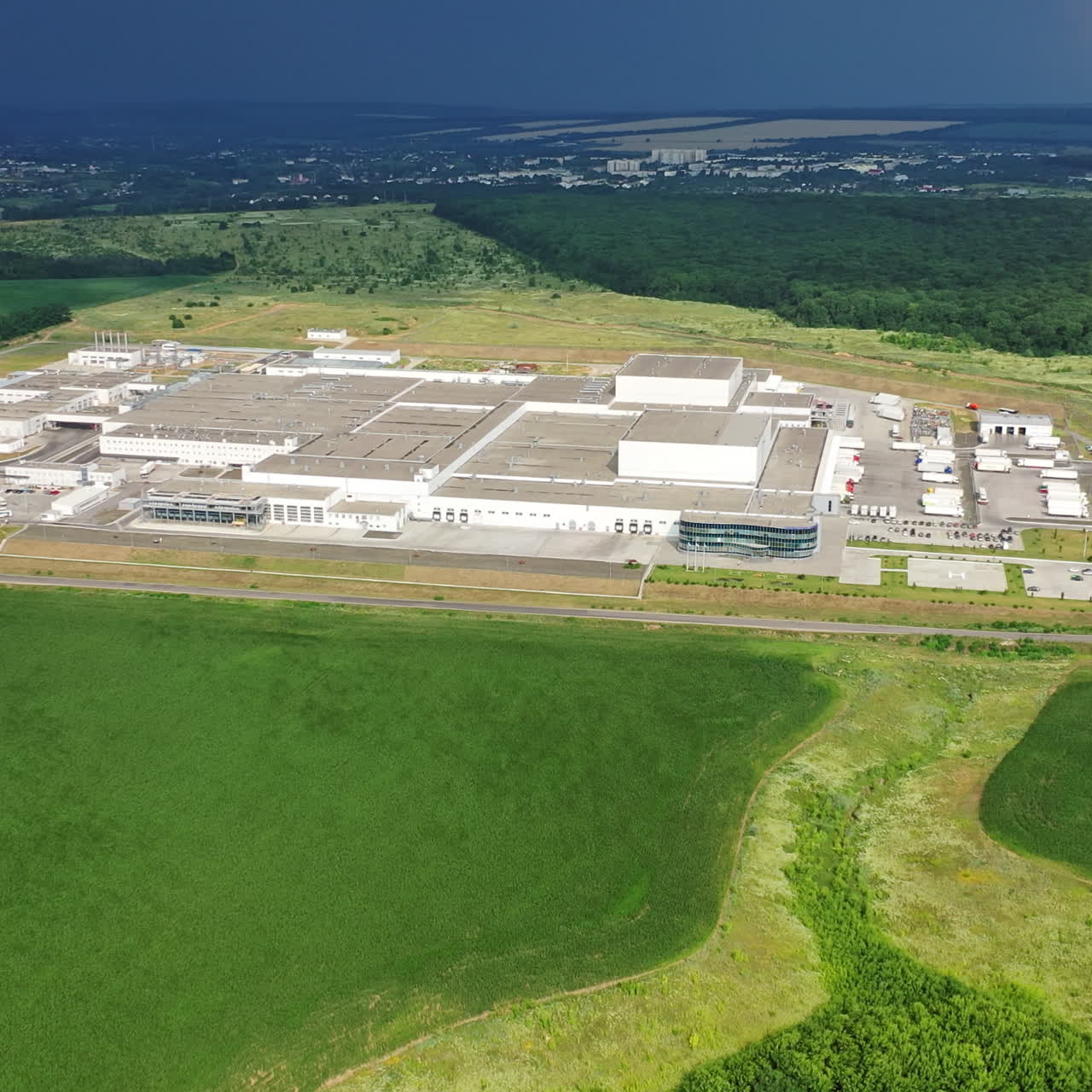 Aerial view from afar on manufacturing complex. Large industrial territory surrounded by green fields and forests in summer. Camera moves down.