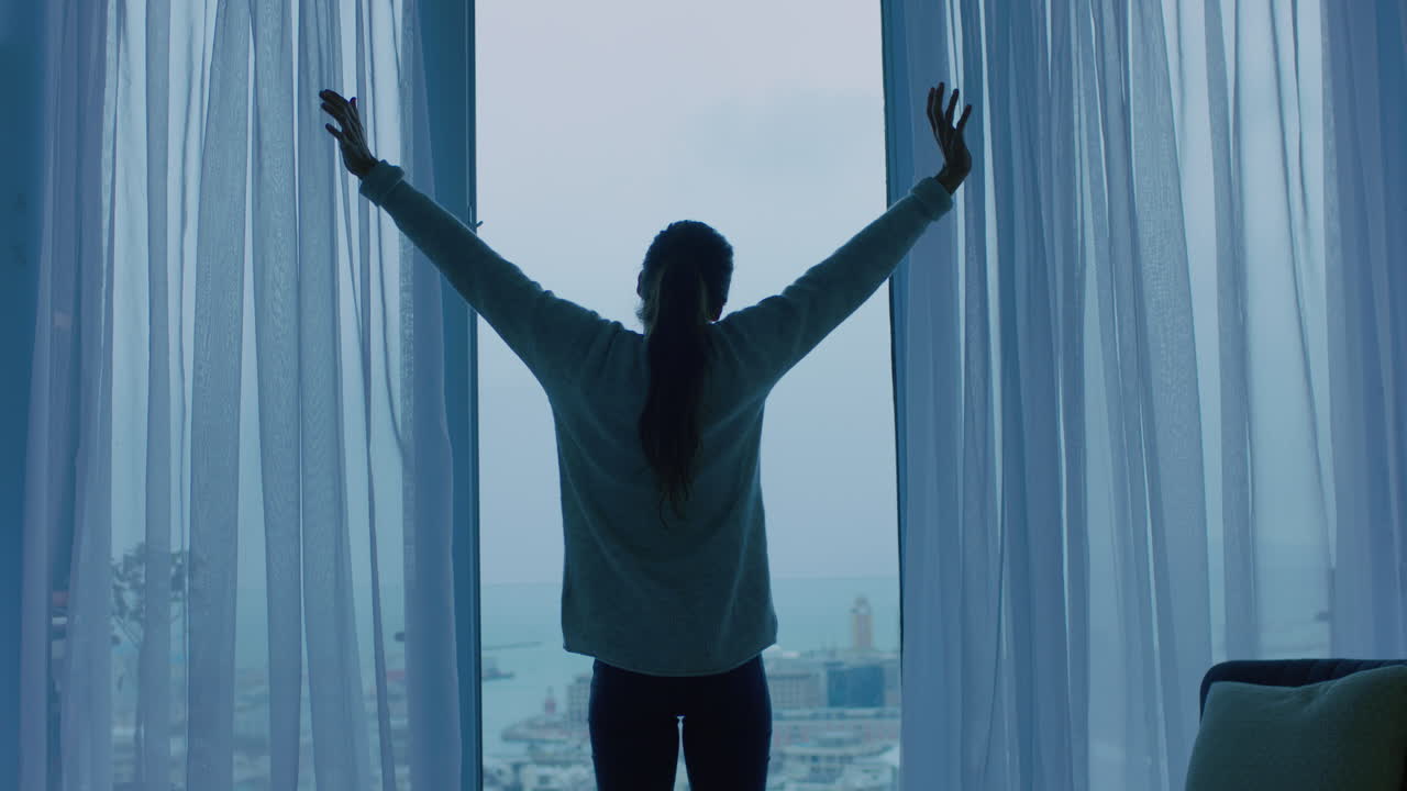 행복한 젊은 여성은 커튼을 열고 창문 밖을 바라보며 신선한 새로운 날을 준비하고 휴식을 취하고 도시의 전망을 즐기고 있습니다.