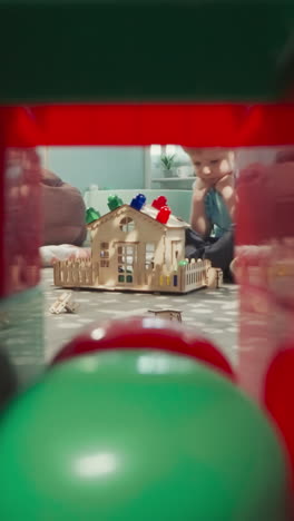 little boy finishes making house from wooden blocks and details. toddler sits on floor playing with toys view through clearance in construction of cubes