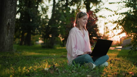 joven con camiseta rosa con gafas en la cabeza, sonriendo cálidamente mientras escribe en su portátil, mira hacia arriba pensativo, luego se vuelve a centrar en su trabajo mientras el sol crea una atmósfera brillante