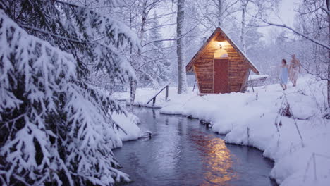 couple in towels walks through snow-covered woods to wooden sauna next to stream