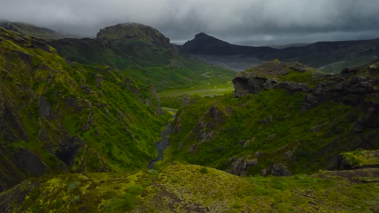 Aerial drone footage flying over green moss and nature covered Iceland volcanic landscape mountains during a cloudy day with large and steep valleys visible in the background. Rocky sharp mountains.