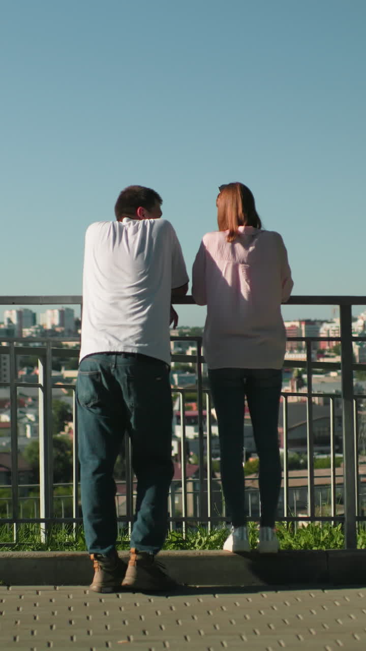 동생들은 철의 레일링에 기대어 그녀의 형제에게 미소 짓는 소녀와 따뜻한 상호 작용을 하고 있습니다. 배경에는 나무와 빛으로 활기찬 도시 풍경이 있습니다.