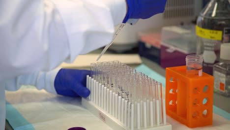 a lab tech in a white coat and blue gloves pipes clear liquid from a pipette into several clear plastic vials