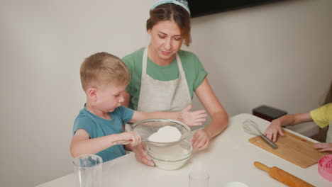 aerial view of young boy sieving flour into bowl held by woman in apron while girl clears cutting board with pink mold and whisk nearby, surrounded by baking tools on clean white kitchen table