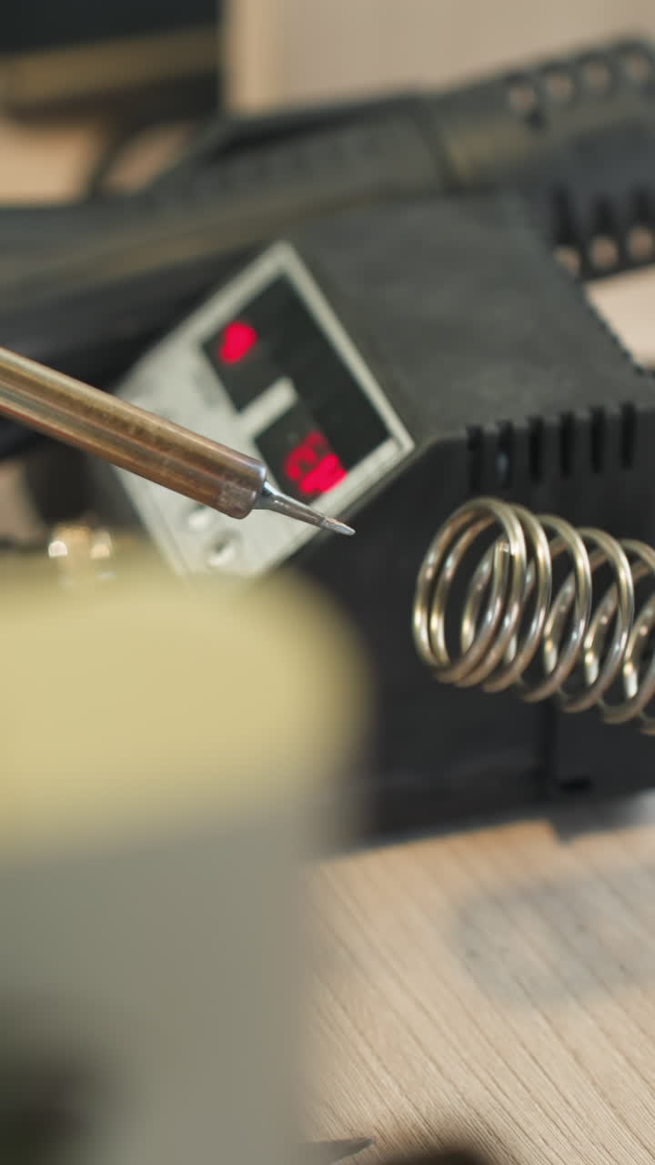 una vista de cerca de una mano con guantes sosteniendo un soldador, colocado en un soporte al lado de una estación de soldadura motorizada, y limpiando la boca de soldadura