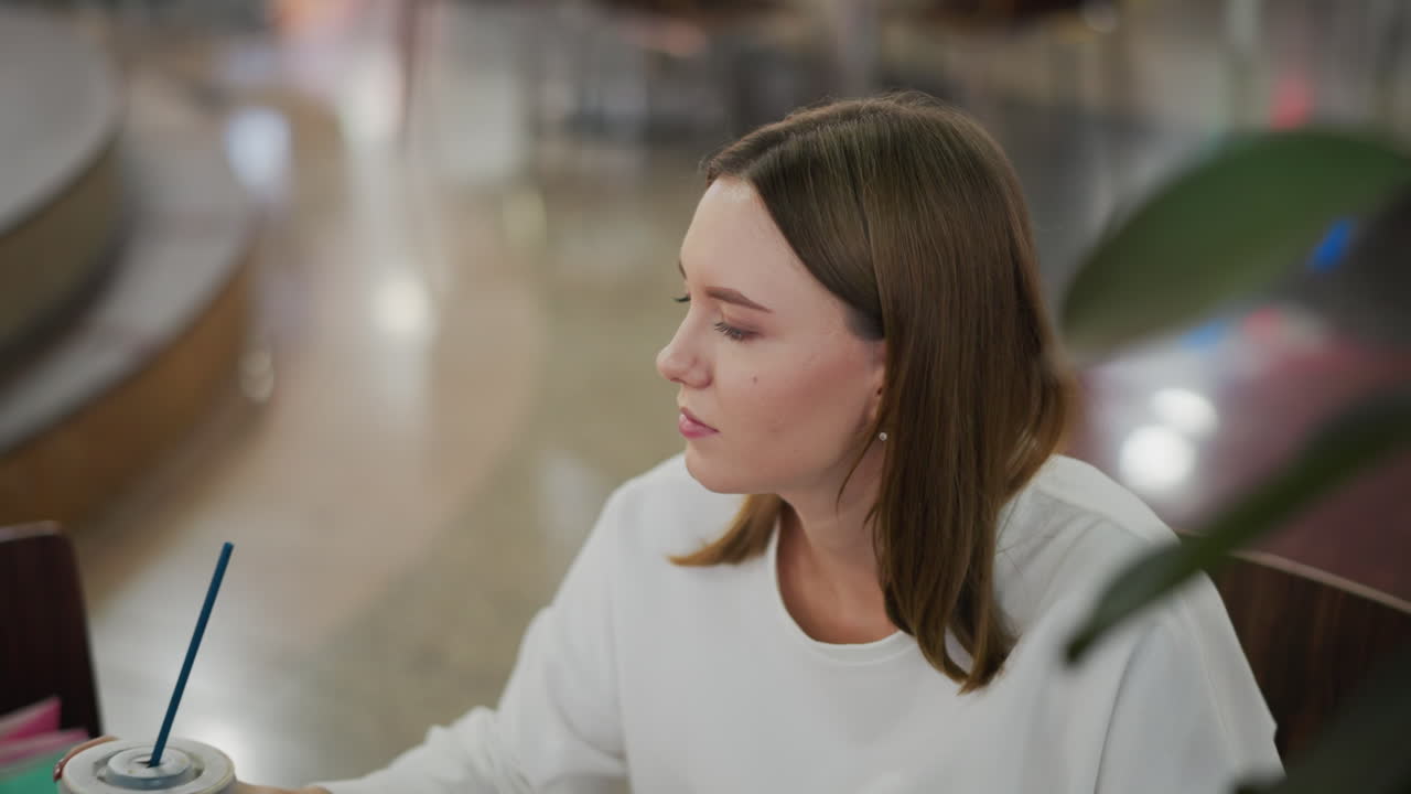 леди в белой рубашке глотает кофе из чашки в расслабленной внутренней обстановке, на заднем плане размыты современные сиденья в ресторане в торговом центре