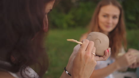 young women apply a pattern a drawing on clay products with the help of tools sticks in a meadow in nature in an open space. a woman decorates a product in close-up.