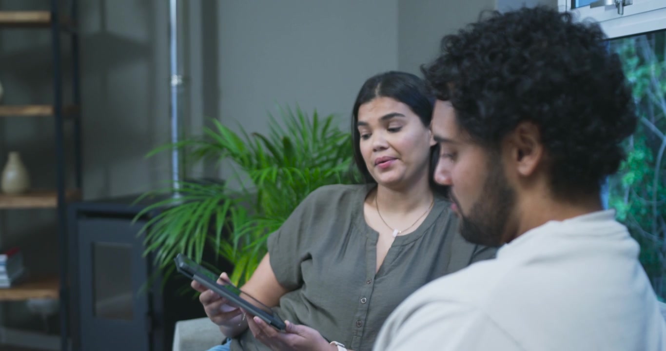 African American couple sitting on couch in home woman sharing phone message man leaning listening
