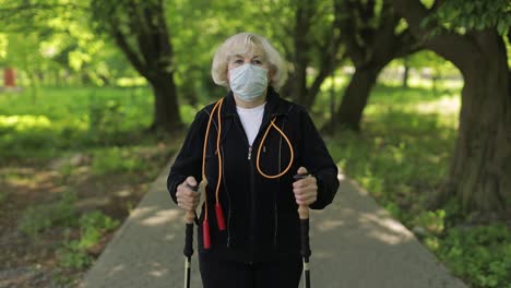 Active-senior-old-woman-in-mask-training-Nordic-walking-in-park-at-quarantine