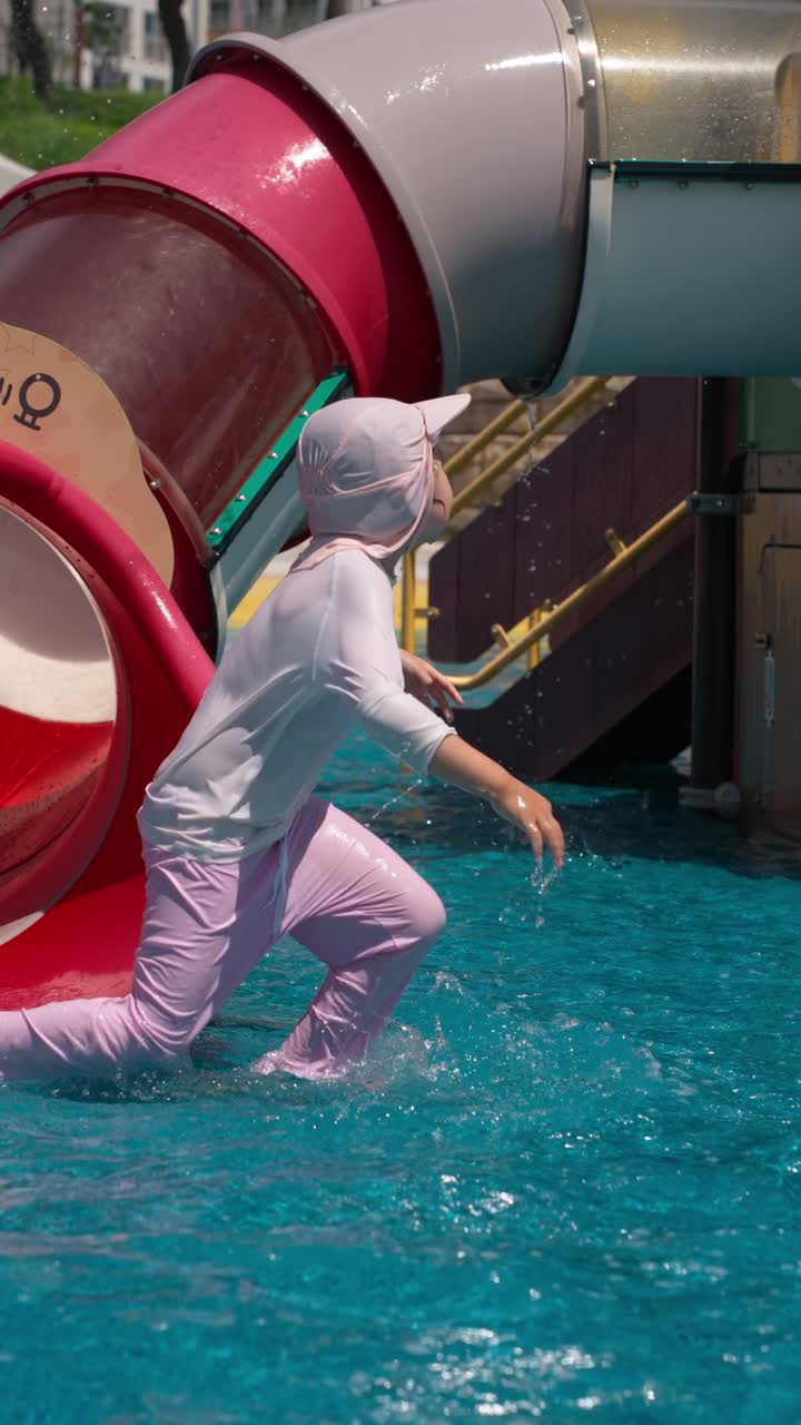 Child slides down tube and laughs while running across shallow splash area in smooth slow motion