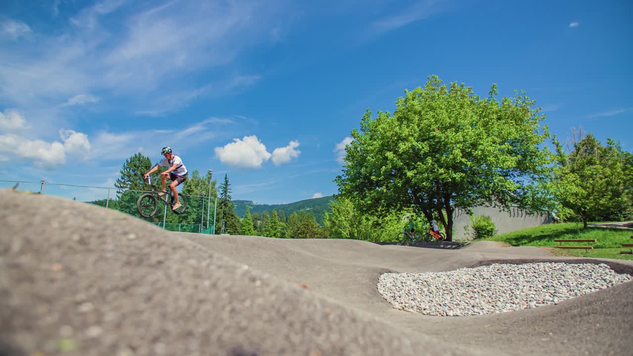un niño montando su bicicleta en una pista de bombeo realizando un truco aéreo, perfil, medio, día soleado, hermoso cielo, nubes blancas, árboles en el fondo