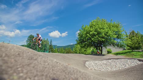 One-boy-riding-his-bicycle-on-a-pump-track-performing-an-aerial-trick,-profile,-medium,-sunny-day,-beautiful-sky,-white-clouds,-trees-in-background