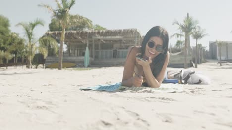 happy hispanic woman in sunglasses lying on beach talking on smartphone, copy space, slow motion