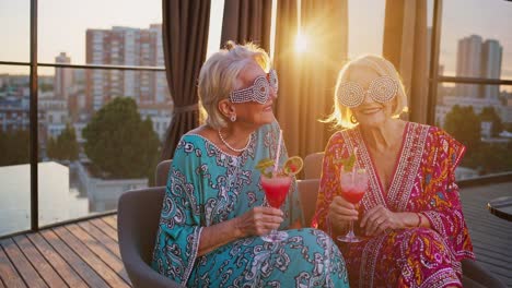 two elegant senior women toasting with cocktails on a rooftop bar, savoring the stunning sunset view over the vibrant city skyline, celebrating friendship and joy in style