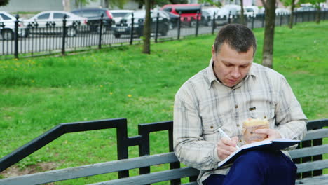 Hombre-Comiendo-Comida-Rápida-En-Un-Banco-Del-Parque-Escribe-Algo-En-Un-Cuaderno