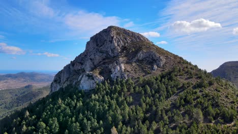 Imágenes-De-Drones-De-La-Ladera-Cubierta-De-árboles-En-Calpe,-España,-Con-Una-Región-Montañosa-Y-Un-Amplio-Paisaje-En-El-Fondo,-Tomadas-En-Un-Día-Soleado-Azul-Brillante-Con-Nubes-Blancas-Esponjosas