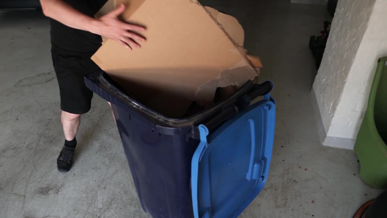 Closer view of a person in casual clothes crushes cardboard boxes on a garage floor and places the pieces into a large blue recycling bin.