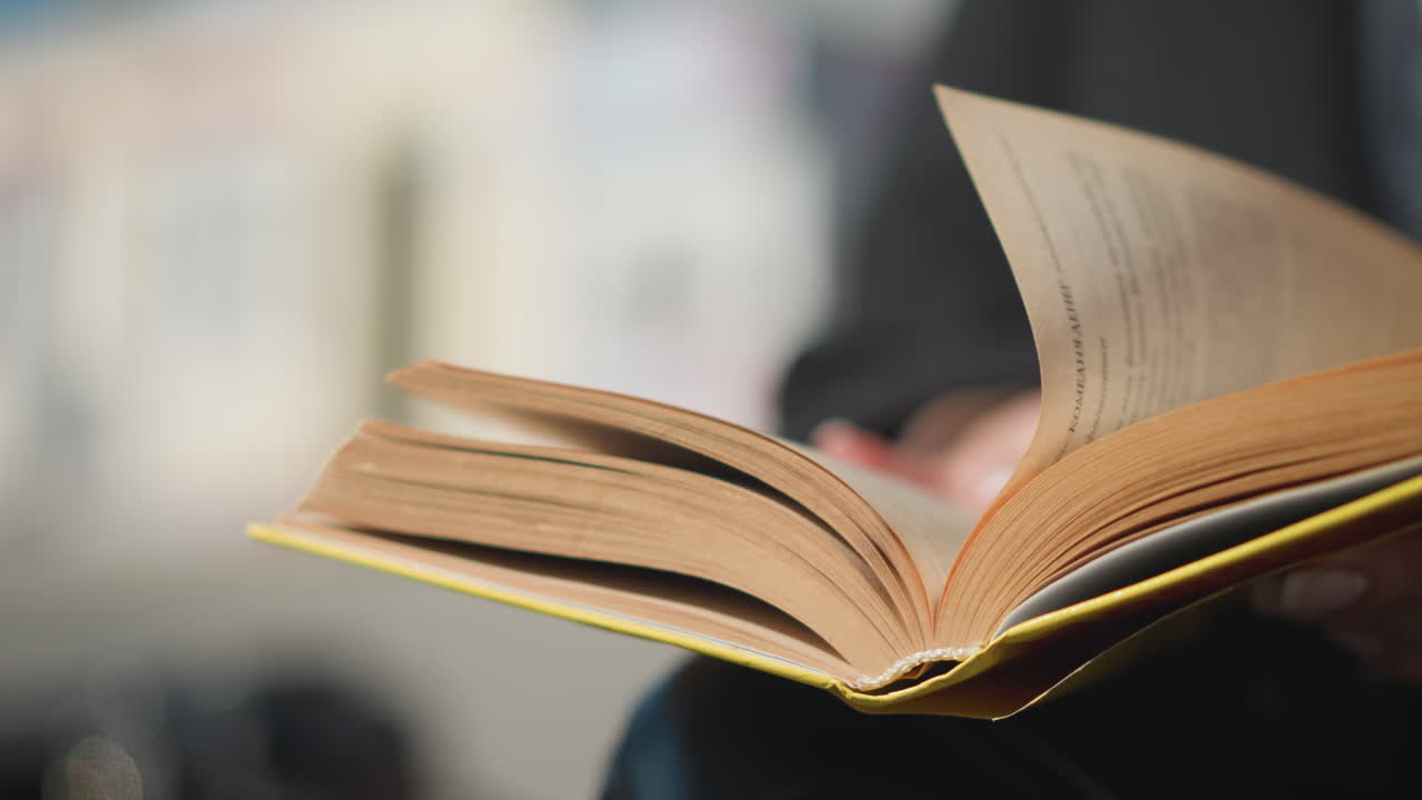 primer plano de una página de un libro siendo volteada por una mano con uñas pulidas borrosas y un anillo, vestido de negro, contra un fondo borroso