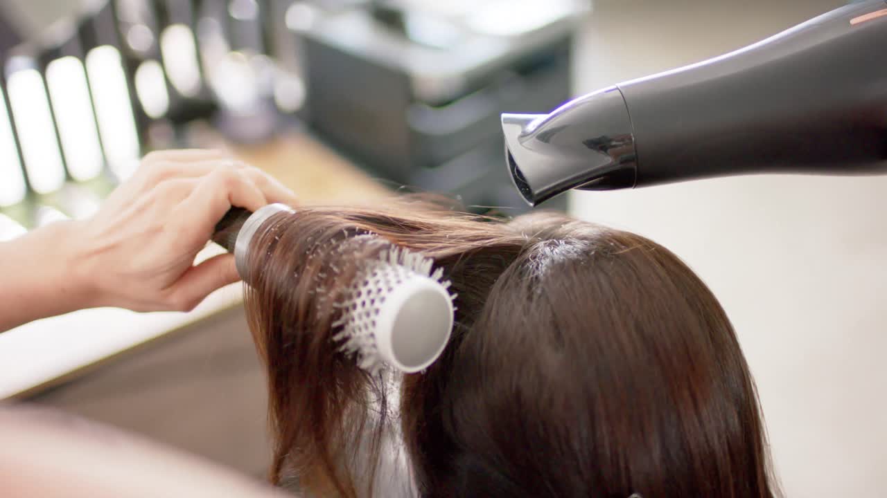 Caucasian female hairdresser styling client's long hair with hairdryer and brush, slow motion