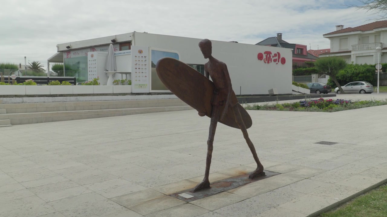 Carrellata del famoso monumento in bronzo del surfista sul lungomare di Salinas, Asturie, Spagna. Mostra la scultura stilizzata, la piazza e l'edificio della scuola di surf locale sotto un cielo luminoso