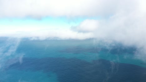 Drone-shot-flying-backwards,-surrounded-by-thin-dancing-clouds-in-the-blue-and-turquoise-sky-with-the-colorful-ocean-below-with-reefs-next-to-Espigao-Amorelo-mountain-in-Madeira