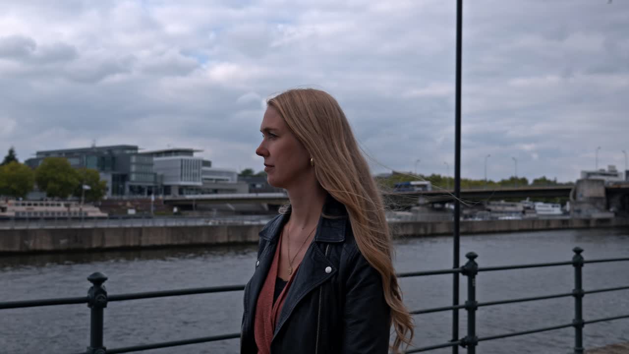 A woman with blonde hair walking along the river promenade on the west bank of the Meuse River (Maas) in Maastricht, Netherlands. The Bridge Sint Servaasbrug (St. Servatius Bridge) in the background