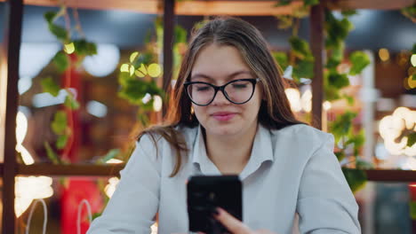 lady engaged with phone, wearing glasses and a warm smile, seated in a decorative indoor setting with glowing lights in the background, lush greenery surrounds her