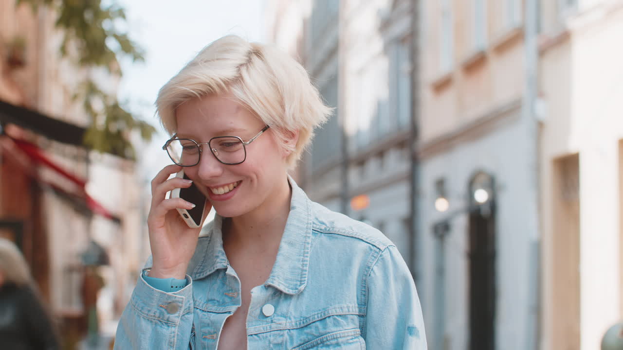 joven rubia caucásica feliz con el cabello corto hablando chismes en el teléfono inteligente en la calle de la ciudad