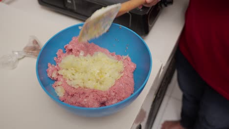 cook adding sautéed onions from a pan to minced beef in a blue mixing bowl