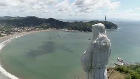 The-Christ-of-mercy-watches-the-beach-of-San-Juan-del-Sur,-aerial-orbit-behind