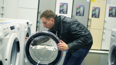 Young-man-is-choosing-a-washing-machine-in-a-store.-He-is-opening-the-doors,-looking-inside