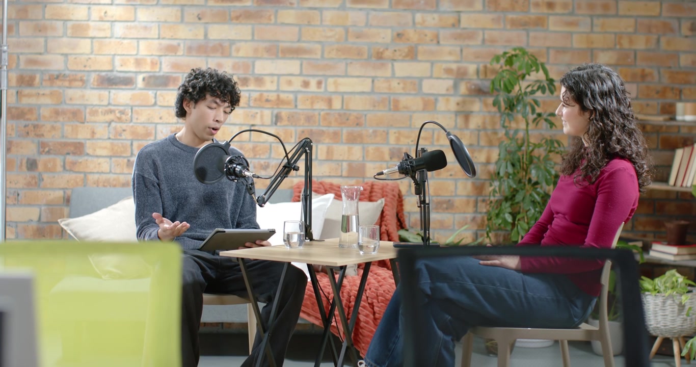 Duo di podcaster afroamericani in studio che leggono da un tablet e parlano al microfono mentre un ospite beve acqua