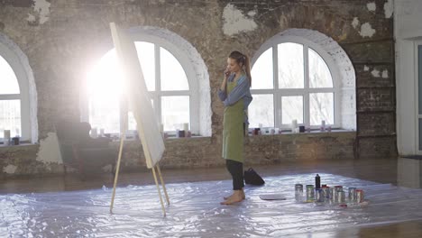 female artist in green apron reflecting in front her artwork in workshop