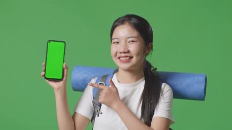 young woman showing mobile phone green screen with backpack and yoga mat