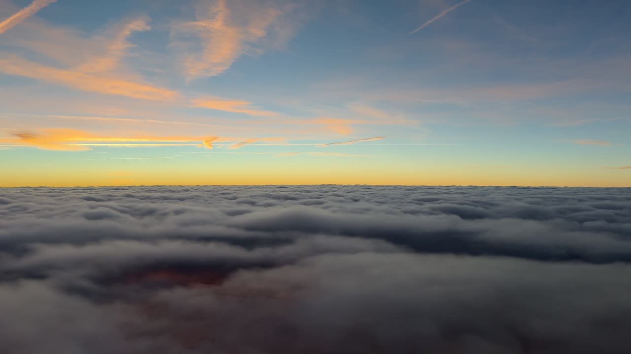 Una coinvolgente visuale in soggettiva dal posto di pilotaggio di un jet che sale sopra un infinito strato di eteree nuvole stratificate grigie, con un orizzonte dorato bagnato dalla luce del sole nascente e scie di condensazione arancioni in alto.