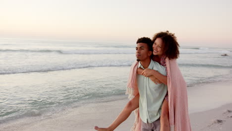 Ein-Gemischtrassiges-Paar-Genießt-Einen-Ruhigen-Strand-Bei-Sonnenuntergang,-Wobei-Die-Frau-Den-Mann-Von-Hinten-Umarmt,-Mit-