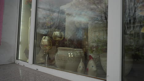 close-up view of a storefront window display with elegant decorative items, including ornate glass goblets, ceramic pots, and plants, with reflection on the glass