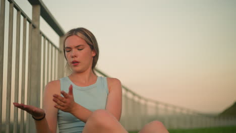 young lady sitting near iron railing cleaning dirt from palm, taking deep breath with eyes closed, evening sunlight gently illuminating face, wearing light blue top, with black hand bangle