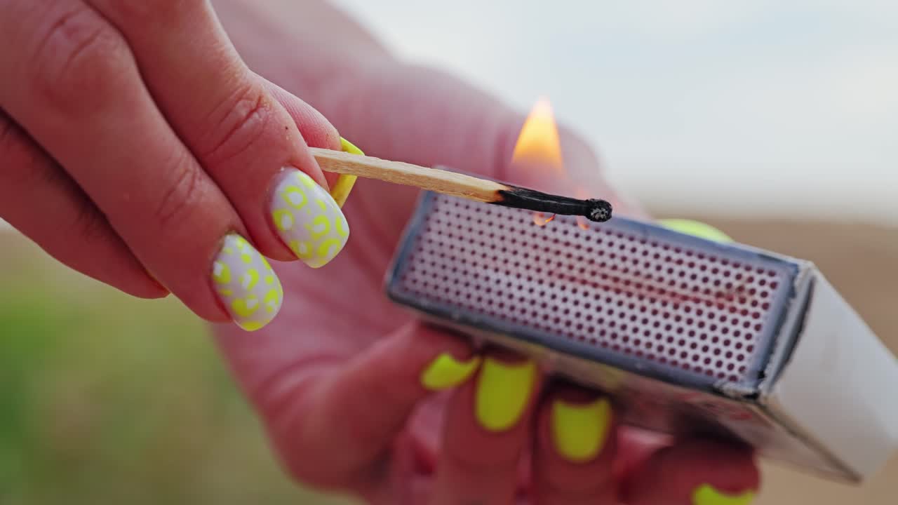 Lit match held by woman outdoors, cooking residue visible beneath bright nails