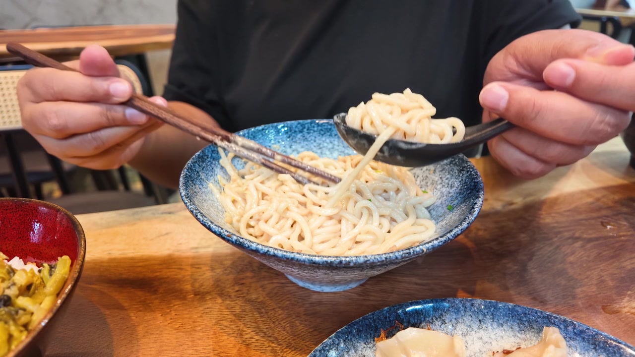 Primo piano delle mani che usano bacchette e un cucchiaio per mangiare noodles taiwanesi a Bangkok.