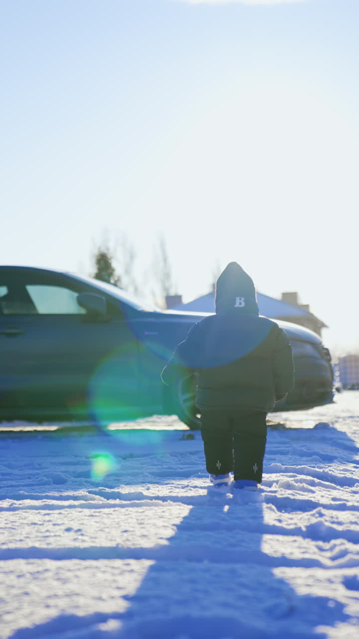 Rear view of a small baby boy wearing warm clothes walking by the snow. Low angle view at the kid going towards the car in sun rays. Vertical video.