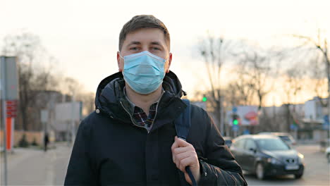 portrait of handsome man in medical mask standing outside at street with backpack and turning face to camera